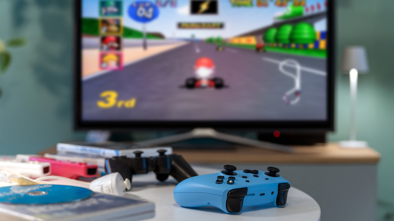 Controllers and game cases on a table with Mario Kart on a flat-screen TV in the background.