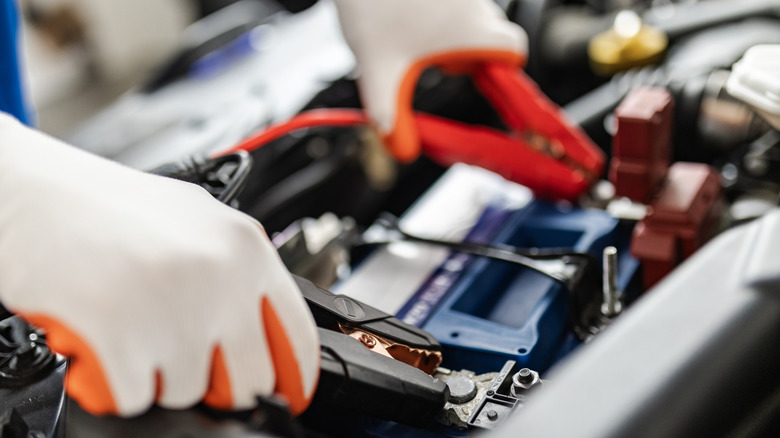 In a well-lit garage, a mechanic in gloves connects jumper cables to a car battery, checking for power issues and preparing for a battery boost to get the vehicle running.