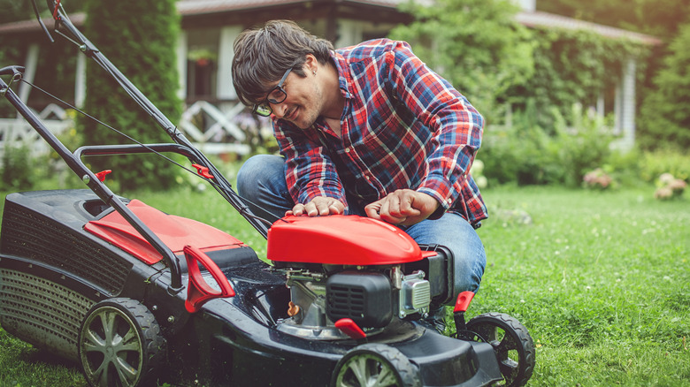 A man wearing eyeglasses, plaid shirt, and denim pants inspecting a lawn mower in a garden.