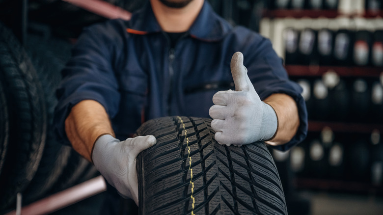 A man in a workshop holding a tire with one hand and showing thumbs up with the other.