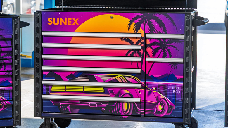 a tool cart with a very 80s looking car under a large sun with silhouetted palm trees in the background