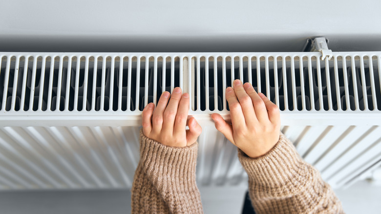 Child wearing brown woolly jacket warming cold hand in front of heating radiator