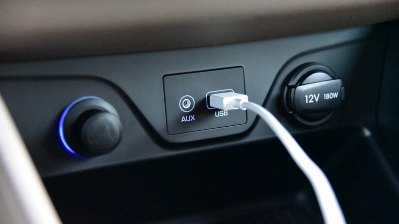 A USB cable plugged into a car's dashboard, AUX port next to it, cigarette lighter to the left, 12V outlet to the right