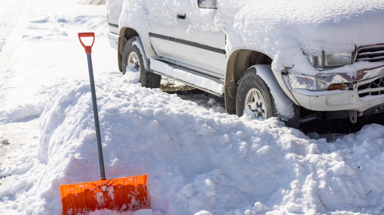 Shovel sticking out of snow heap with car covered in snow behind it