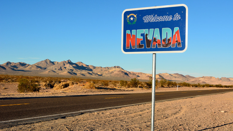 A blue "Welcome to Nevada" traffic sign with desert sands, hills, and a remote road in the background.