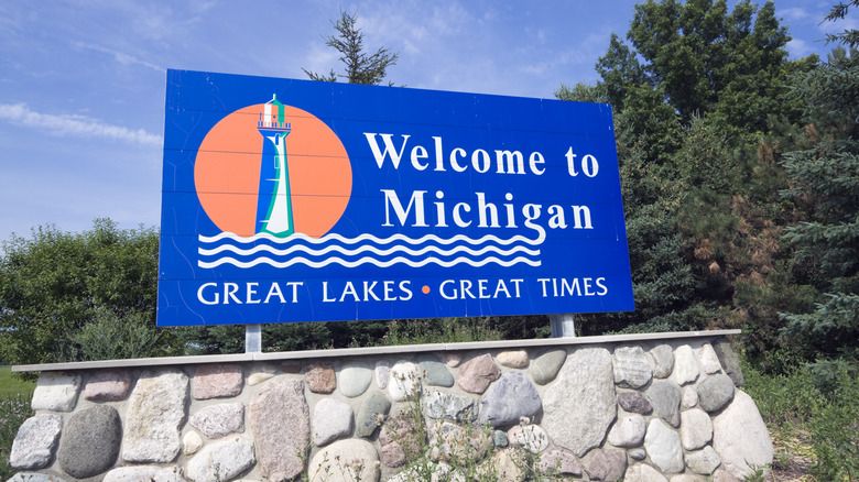 Blue 'Welcome to Michigan' sign atop a rock wall with trees and blue sky the background.