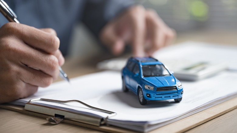 Person writing on a paper form with blue toy car on desk.