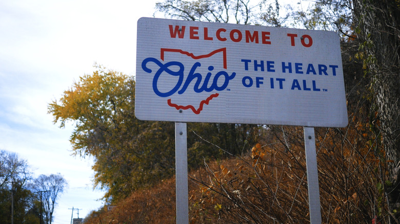 A white road sign reading "Welcome to Ohio," with autumn trees and grass in the background.