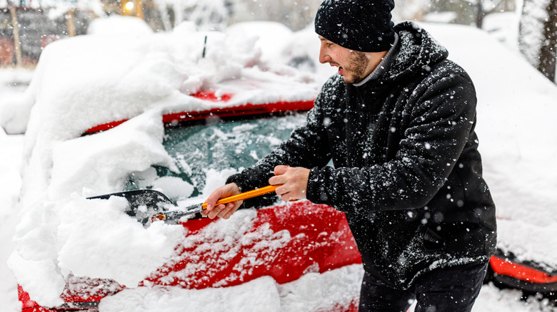Man is Cleaning Snow from a Car with a Snow Shovel. Man Outdoors is Cleaning Snow on his Car Windshield.