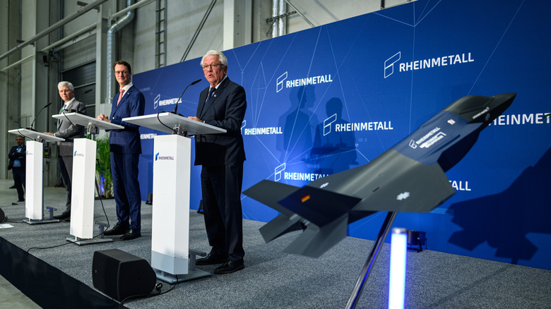 Three people on stage at a news conference, with a model of a German-built F-35 fighter in the foreground