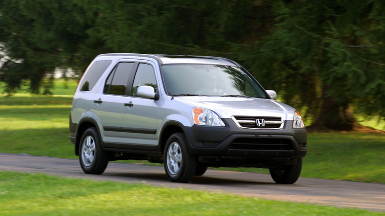 A silver second generation Honda CR-V driving on a paved roadway through a park-like setting.