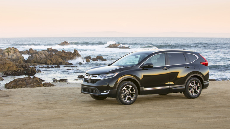 A black fifth generation Honda CR-V parked on a beach, with waves hitting the rocks beyond.