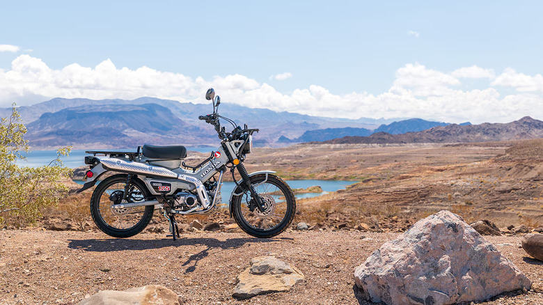 A silver Honda Trail 125 parked upright against a scenic natural backdrop with a lake in the back.