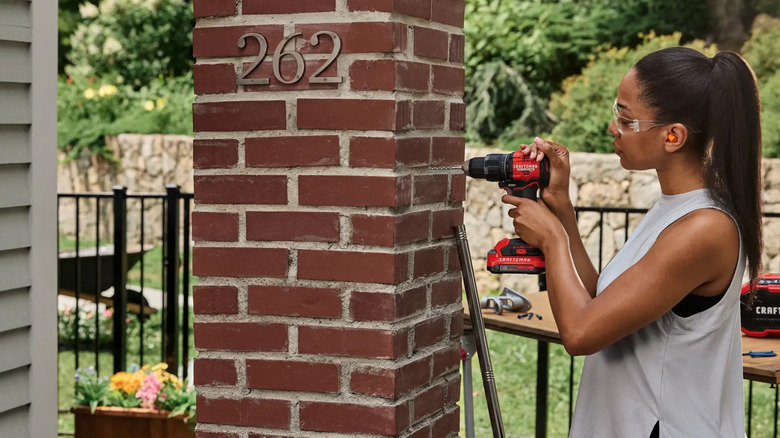 A woman drilling a hole into a brick column on a house.
