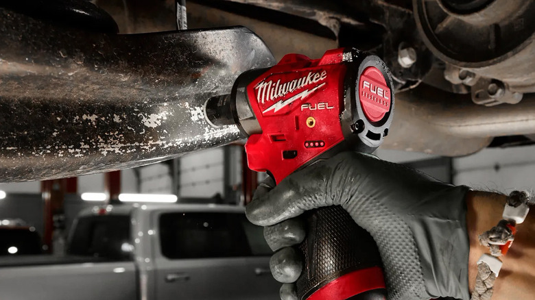 A person using a Milwaukee impact wrench to loosen a bolt on a car frame