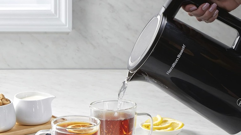 Hand pouring water from a black Hamilton Beach electric kettle into a glass mug