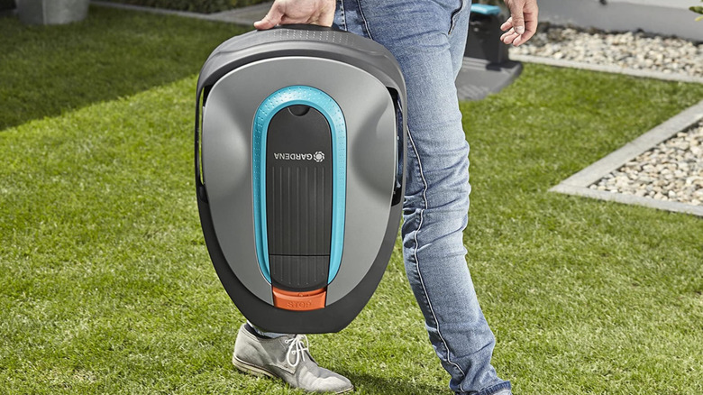 A person carrying a Gardena robot mower by hand in a yard.
