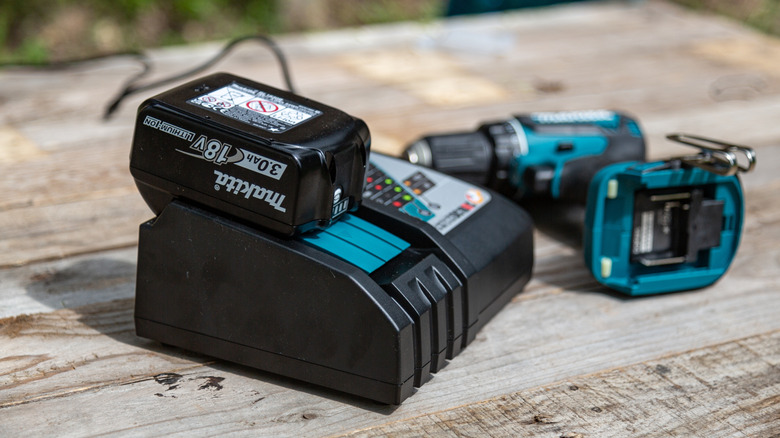 A Makita battery in charge next to a Makita drill missing a battery on a wooden table.
