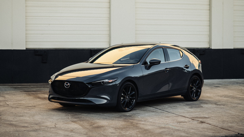 Gray 2025 Mazda3 hatchback parked in front of warehouse, front 3/4 view