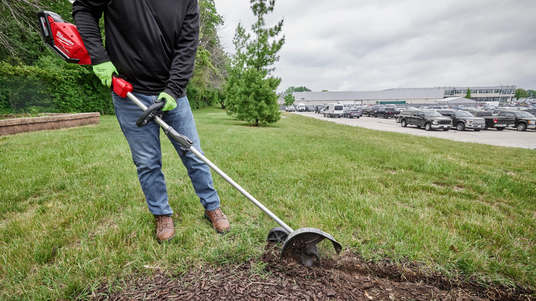 A person uses a Quik-Lok bed redefiner to cut a V-shaped trench between the grass and a garden bed.