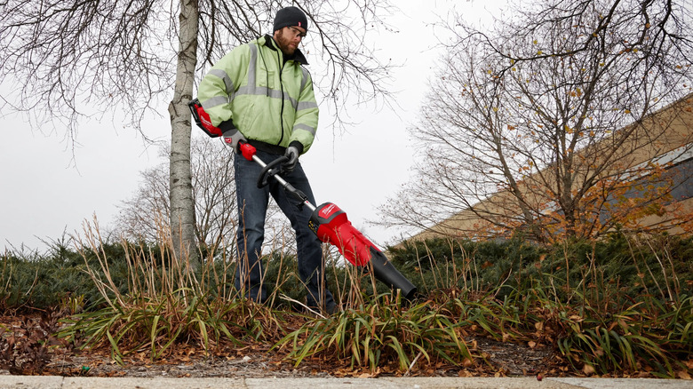 A man in a hat and safety glasses uses a Quik-Lok blower to remove dead leaves from a garden bed during the cold season.