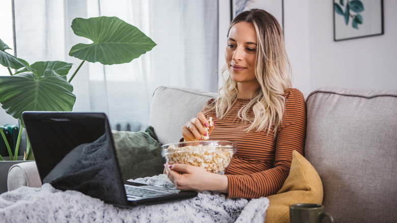 woman on a couch eating popcorn and looking at laptop