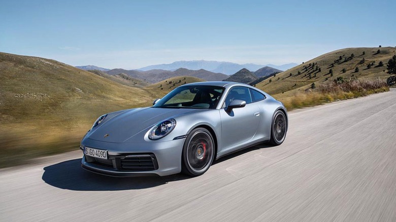 A silver Porsche 911 model from the 992.1 generation moving at speed down a road in a sandy area.