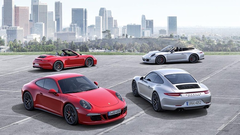 Four different Porsche Carrera GTS models parked on a paved surface with a city and forest in the backdrop.