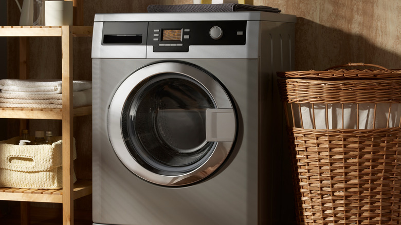 A silver front-load washing machine positioned next to a wooden shelving unit and a wicker laundry basket.