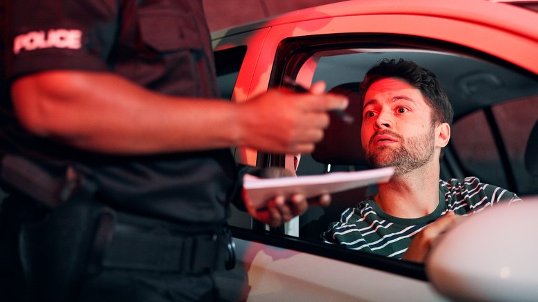 A police officer holding a pen and pad paper while standing close to the side of a car with a male driver looking out of the window.