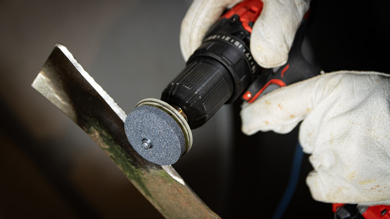 Gloved hands operating a power drill to sharpen a lawn mower blade