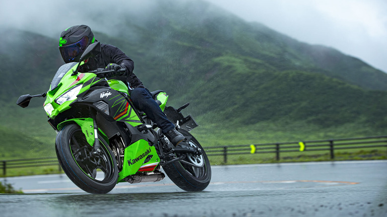Rider leaning into a turn on a bright green Kawasaki Ninja ZX-25R sportbike in wet conditions, with misty green mountains and a wet road surface in the background.