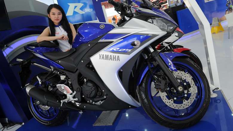 Yamaha R25 sportbike in blue and silver on display at an indoor showroom, with a promotional model posing beside the motorcycle and Yamaha branding visible in the background.