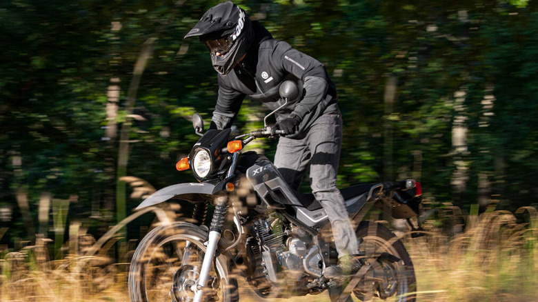 Motorcyclist wearing full gear and a black helmet riding a Yamaha XT250 off-road through tall grass, with motion blur in the background indicating speed and movement.