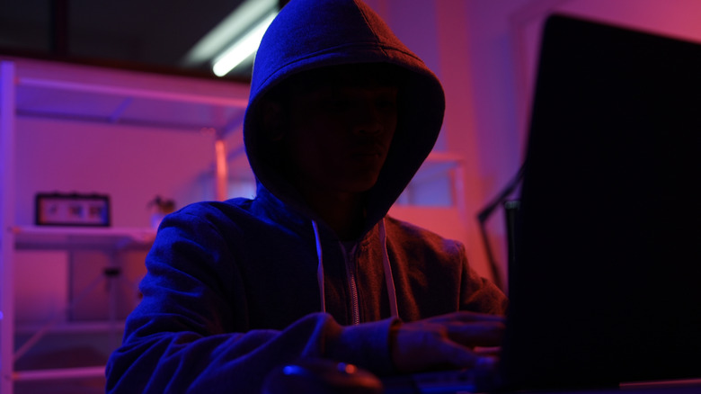 A masked and hooded figure using a laptop.