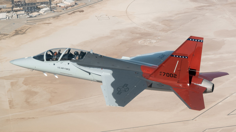 T-7A Red Hawk jet seen from above left flying above a desert airbase