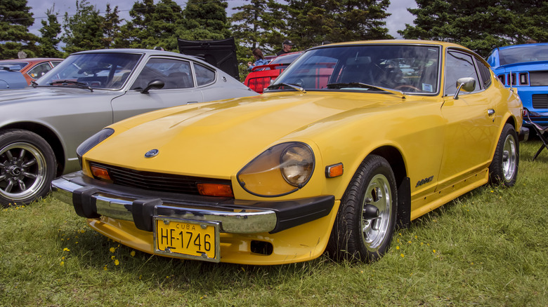 A yellow 1970s Datsun 280Z parked on a lawn with others cars around it.