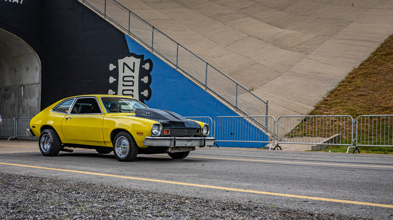 A 1970s yellow modified Ford Pinto being driven on an urban public road.