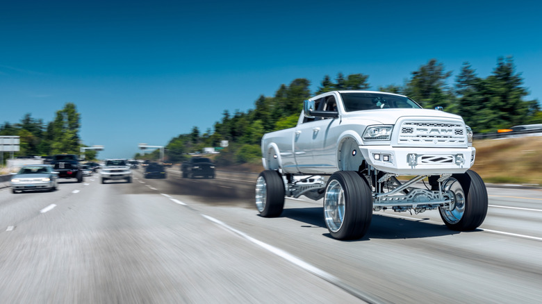 A massive lifted all-white diesel Ram truck driving on a highway under blue skies.