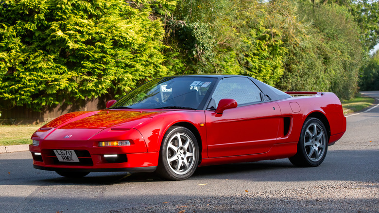 A front three-quarter view of a parked red early-gen Honda NSX supercar with trees in the background.
