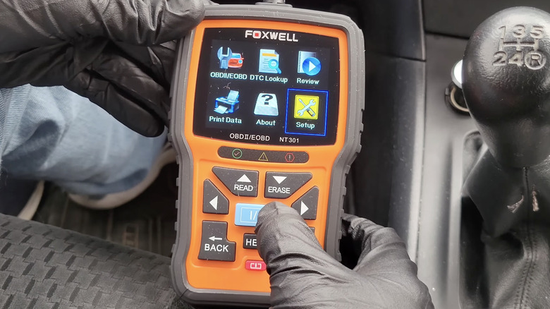 A person wearing black gloves holding a Foxwell OBD-II scanner near a car's gear shifter.
