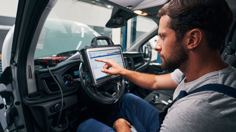 A mechanic sitting inside a vehicle with the steering wheel holding a tablet showcasing car diagnostics software.