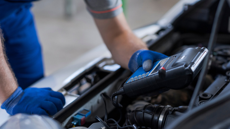 A mechanic in blue gloves holding a scanner above a car's engine bay.