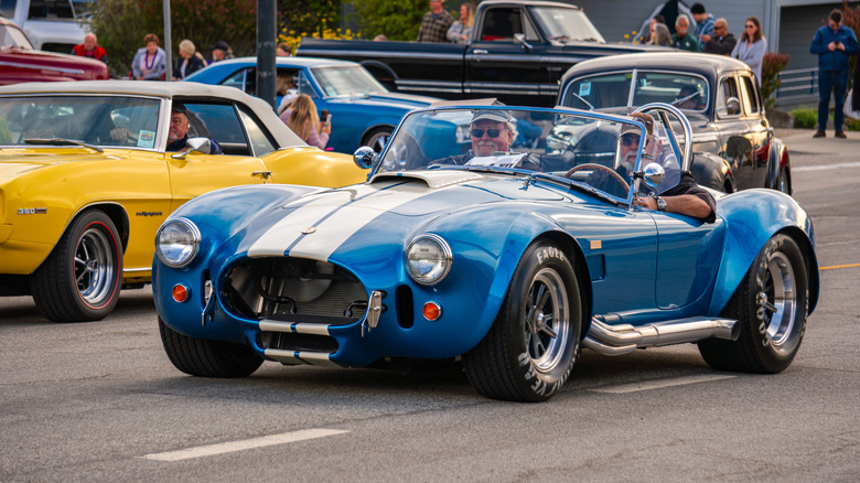 A blue 427 1960s Shelby Cobra driven on a road alongside other vintage American vehicles.