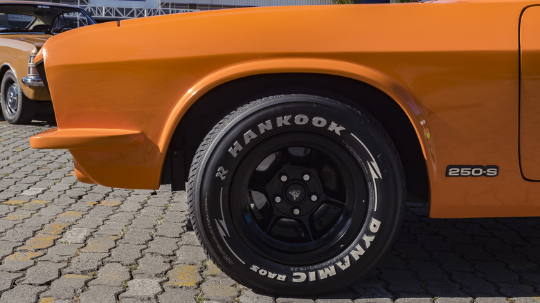 A front-end side view of a restored old 1970s orange classic car with black wheels and Hankook tire writing.