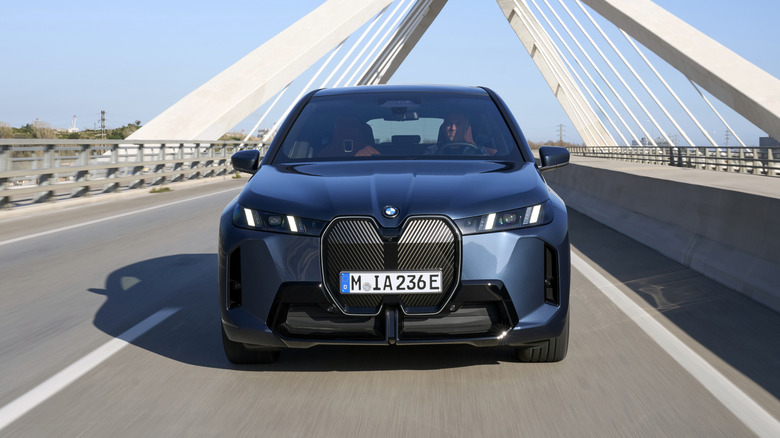A front-end view of a blue BMW iX being driven on a bridge with blue skies in the background.