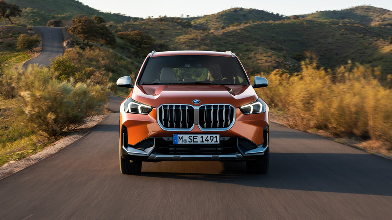 A front-end view of a metallic orange 2023 BMW X1 driving on a rural road.