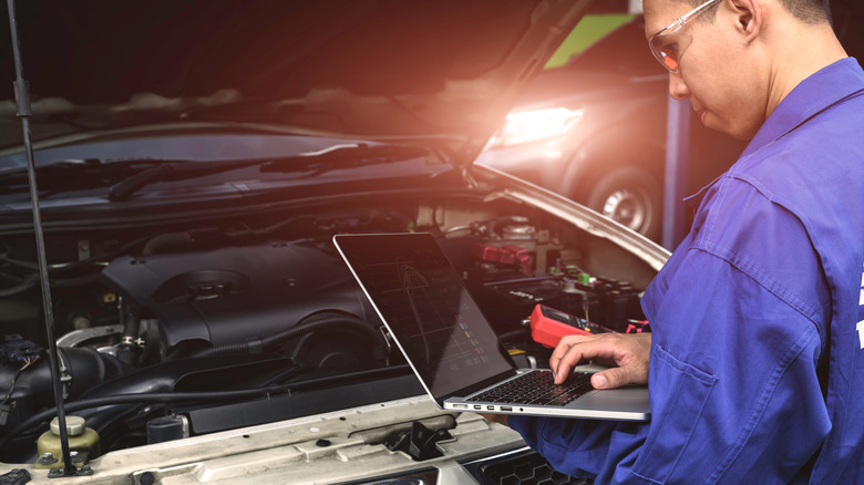 A mechanic with a laptop standing over a car's open hood.