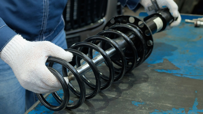 A mechanic wearing white gloves holding a black shock absorber on a work bench.