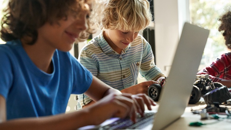 Middle school-aged boy on a laptop while another fiddles with an RC car